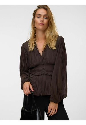 Femme aux cheveux blonds porte un chemisier plissé marron foncé et un pantalon noir, tenant un sac à main en cuir noir sur fond blanc.