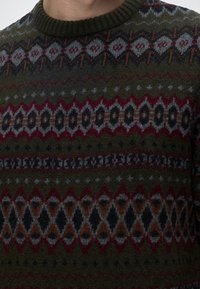 Gros plan d'un pull en laine avec un motif géométrique multicolore dans des tons verts, rouges, gris, noirs et bruns.