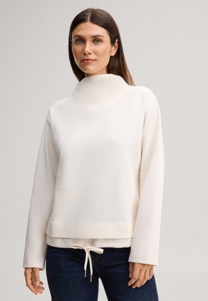 Femme aux cheveux bruns raides portant un pull crème à col montant, manches longues avec un cordon de serrage à l'ourlet et un jean bleu foncé.