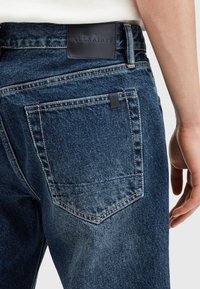 Jeans in denim blu con una toppa in pelle etichettata "ALLSAINTS", cuciture color marrone chiaro e una tasca posteriore visibile nell'immagine.