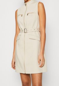 Belted sleeveless dress in beige fabric, featuring gold zipper details, two chest pockets, and a high neckline. Smooth texture throughout.