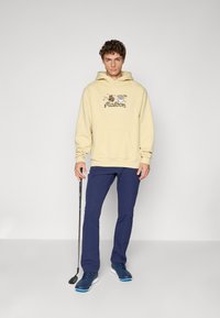 Beige hoodie with a graphic design featuring cartoon animals and text, paired with navy pants and blue athletic shoes. Holding a golf club.