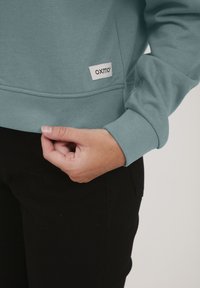 Turkos sweatshirts med rund halsringning och ribbade muddar. Har en liten vit etikett med "OXMO" på vänster nederkant. Svarta byxor under.