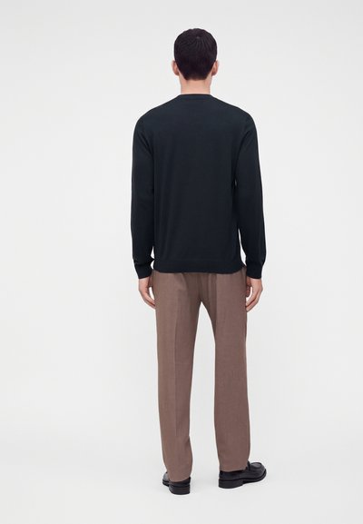 Homme vu de dos portant une chemise noire à manches longues, un pantalon marron et des chaussures habillées noires sur fond blanc.