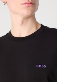 Fekete pamut póló bordázott kerek nyakkal és kis lila "BOSS" logóval a bal mellkas területén. Sima textúra, laza dizájn.