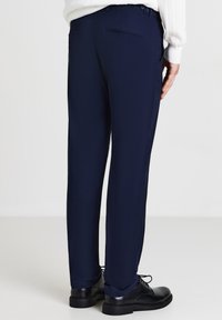 Homme portant un pantalon habillé bleu marine et des chaussures en cuir noir, se tenant contre un fond clair, visible des genoux vers le bas.