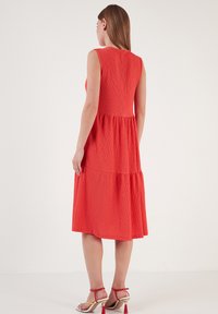 Rotes, ärmelloses Kleid aus strukturiertem Stoff, mit gestuftem Design und knielangem Saum. Getragen mit Riemchensandalen mit Blockabsatz.