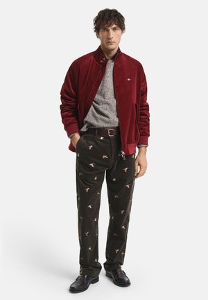 Mann in einer burgunderfarbenen Jacke, grauem Pullover, dunkel gemusterten Cordhosen mit Vögeln, schwarzem Gürtel und dunklen Lederschuhen, der mit den Händen in den Taschen steht.