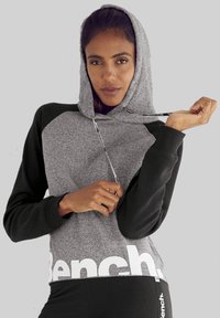 Felpa grigia e nera con tasca a marsupio frontale, cappuccio con coulisse e grande logo bianco "Bench" in basso.