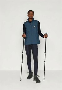 Μπλε γιλέκο με φερμουάρ μπροστά πάνω από ένα μαύρο μακρυμάνικο πουκάμισο, συνδυασμένο με μαύρα κολάν και ορειβατικά μποτάκια. Κρατάει μπαστούνια πεζοπορίας, σε ουδέτερο φόντο.