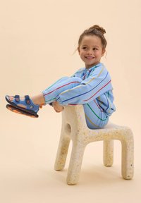 Tuta leggero blu chiaro con strisce multicolori, abbinata a sandali blu con accenti arancioni, seduta su una sedia bianca testurizzata.