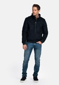 Marineblauwe gewatteerde jas met opstaande kraag en ritssluiting, gecombineerd met blauwe jeans en zwarte schoenen. Beschikt over zijzakken en geribbelde manchetten.