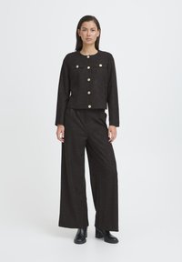 Donna che indossa una giacca nera testurizzata con bottoni dorati e pantaloni a gamba larga abbinati, abbinati a stivaletti neri, in piedi.