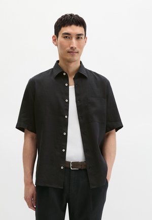 Mann trägt ein kurzärmliges schwarzes Hemd mit Knöpfen über einem weißen Tanktop, schwarze Hose und braunen Gürtel, steht vor weißem Hintergrund.