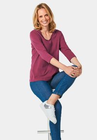 Bordeauxfarbener Langarm-Oberteil mit V-Ausschnitt, kombiniert mit blauen Jeans und weißen Sneakers mit silbernen Akzenten. Sitzende Pose, neutraler Hintergrund.