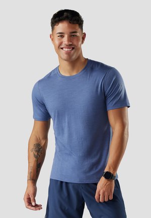 Junger Mann, der lächelt und ein blaues kurzärmeliges Hemd, dunkelblaue Shorts, eine schwarze Smartwatch und eine Sonnenbrille auf dem Kopf trägt, mit einem Rosen-Tattoo auf seinem linken Unterarm.