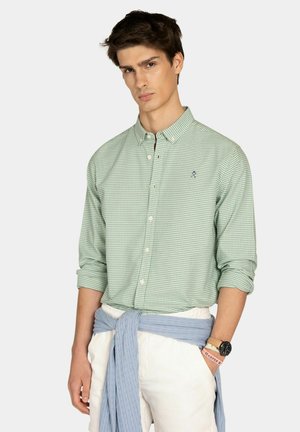 Joven con camisa de cuadros verde abotonada, pantalones blancos y un suéter azul claro atado a la cintura, posando contra un fondo liso.