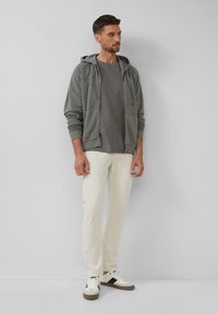 Grau zip-up Hoodie über einem grauen gerippten T-Shirt, kombiniert mit cremefarbenen Hosen und weißen Sneakers mit schwarzen Akzenten.