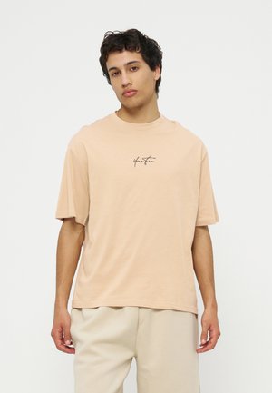 Νέος άνδρας με κοντά σγουρά μαλλιά φοράει μια μπεζ φαρδιά μπλούζα με μαύρο κείμενο και ανοιχτό μπεζ παντελόνια, στέκεται μπροστά σε λευκό φόντο.