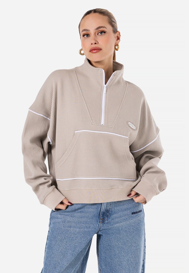 Beige kvart-lukket genser i teksturert stoff, med hvite detaljer, en frontlomme, og en avslappet passform. Kombinert med lyseblå jeans.