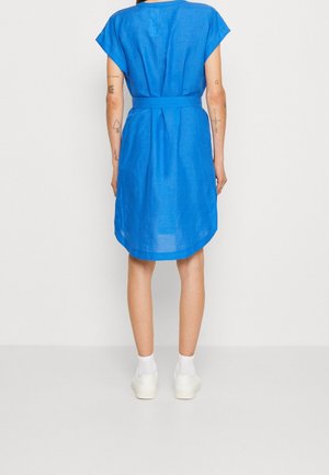 Personne portant une robe bleue à manches courtes avec une ceinture à la taille, des chaussettes blanches et des baskets blanches, debout de dos devant un fond uni.