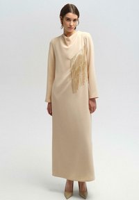Beige lange mouw jurk met gedrapeerde halslijn, voorzien van een diagonale franjerand in goud. Gladde textuur, vloerlange ontwerp, gedragen met beige hakken.