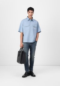 Lyseblå kortermet skjorte med knapper og to brystlommer, kombinert med mørke jeans og svarte sko, og bærer en svart totebag.