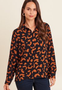 Frau mit einem langärmeligen schwarzen Shirt mit einem orangefarbenen Leopardenmuster und dunkelblauen Jeans, die vor einem beigen Hintergrund steht.