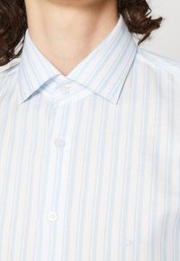 Camisa de vestir a rayas azul claro y blanca con cuello abotonado y tapeta frontal. Presenta un sutil logo "CK" bordado en el lado inferior.