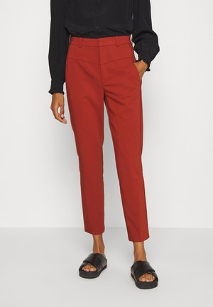 Pantalon sur mesure rouge rouille avec poches latérales, porté avec un chemisier noir à manches longues et des sandales noires sans bride, sur un fond uni.