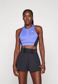 Sportliches Crop-Top in Helllila mit schwarzen Seiten, das eine eng anliegende Passform hat und das Adidas-Logo zeigt. Dazu weite schwarze Sportshorts.