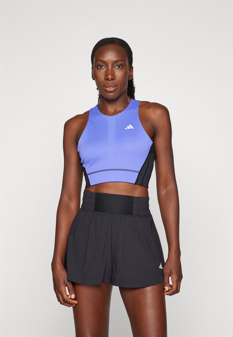 Sportliches Crop-Top in Helllila mit schwarzen Seiten, das eine eng anliegende Passform hat und das Adidas-Logo zeigt. Dazu weite schwarze Sportshorts.