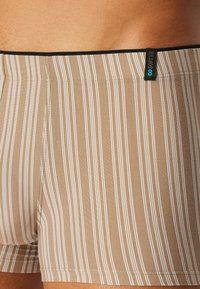 Beżowe kąpielówki z pionowymi białymi paskami, elastycznym paskiem w talii i haftowanymi detalami. Metka na pasku wskazuje cechy marki.