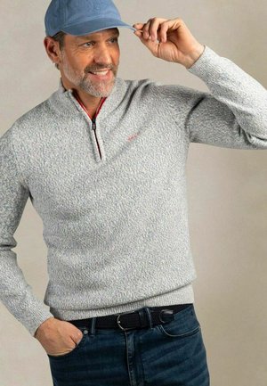 Middle-aged man adjusting light blue cap, wearing grey textured quarter-zip sweater and dark blue jeans with black belt against neutral background.