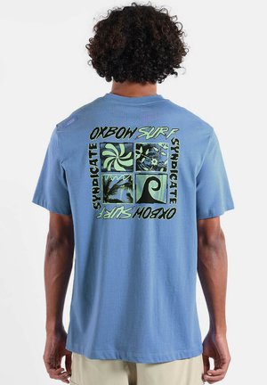 Homme portant un t-shirt bleu avec un graphique à thème surf et le texte "OXBOW SURF SYNDICATE" au dos, debout devant un fond uni.