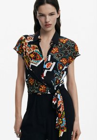 Collared blouse featuring vibrant floral and geometric patterns in red, orange, blue, and green, with a wrap design and tie accent at the waist.
