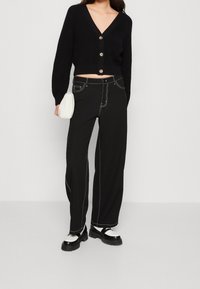 Zwarte cropped cardigan met een V-hals en gouden knopen, gecombineerd met zwarte broek met wijde pijpen en witte stiknaden, en robuuste zwarte en zilveren schoenen.