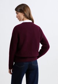 Burgundowy sweter dzianinowy z okrągłym dekoltem i luźnym krojem. Posiada ściągacze na mankietach i u dołu, noszony na ciemnoniebieskich dżinsach. Gładka tekstura.