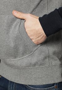 Szary sweter z kieszenią na przodzie, o puszystej fakturze i ściągaczach na mankietach. Widoczna ręka spoczywająca w kieszeni.