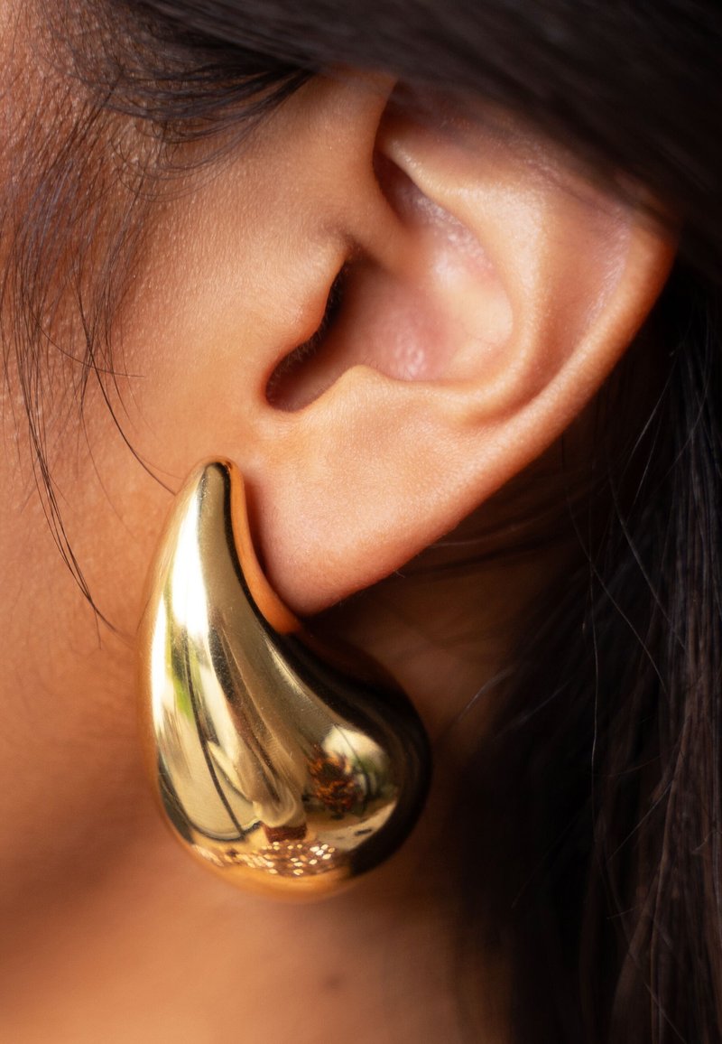 Boucle d'oreille en or en forme de goutte avec une surface lisse et brillante, attachée à un lobe, mettant en valeur sa forme courbée et sa qualité réfléchissante.