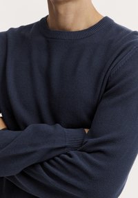 Pull en tricot bleu marine avec une surface texturée, un col rond et des poignets côtelés. Présente une coupe ample avec des coutures d'épaule tombantes.