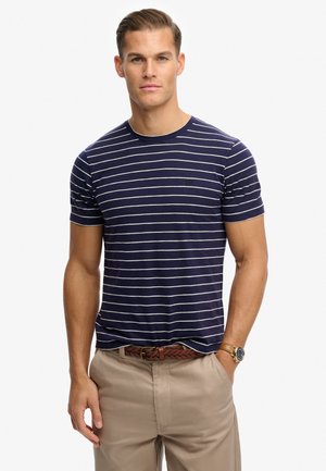 Uomo con camicia a righe blu navy e bianca a maniche corte, pantaloni beige, cintura marrone intrecciata e orologio da polso, in piedi con una mano in tasca.