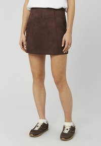 Mini-jupe en daim marron, coupe ajustée, texture lisse, associée à des baskets marron foncé avec des lacets et des accents blancs, portée avec un haut blanc.