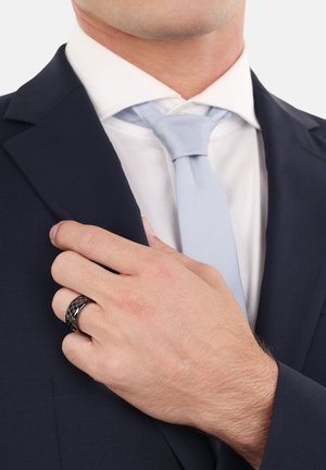 Schwarzer Ring mit einem strukturierten Muster an einer Hand. Die Hand richtet eine dunkelblaue Sakkokur über einem weißen Hemd und einer hellblauen Krawatte.