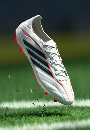 Weißer Fußballschuh mit schwarzen Streifen, roten Akzenten und pinken Stollen; strukturiertes Lederfinish; erhöhtes Nähen auf dem Obermaterial sichtbar.