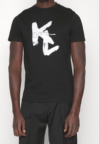 Black cotton t-shirt featuring a large white graphic with "K" and "C" letters. Details include brand name and address printed below the graphic.