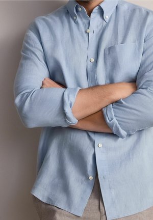 Person trägt ein hellblaues Hemd mit Knöpfen, die Ärmel sind hochgekrempelt, steht mit verschränkten Armen vor einem schlichten Hintergrund.