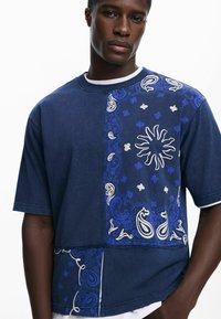 Marineblå kortærmet T-shirt med paisley-mønster i blå og hvid, med et solgrafik og kontrasterende teksturerede sektioner.