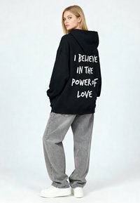 Črni oversized pulover s kapuco z belim napisom na hrbtu. Kombiniran z sivimi kavbojkami in belimi supergami. Gladek material in sproščen kroj.