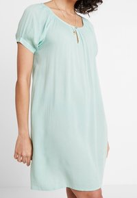 Robe vert menthe clair avec de courtes manches bouffantes, encolure froncée et détail en trou de serrure. Tissu doux et coupe décontractée. Style minimaliste.
